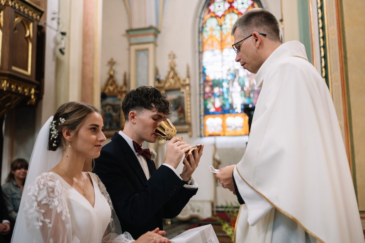 Priest offers communion cup to young groom and bride during Catholic wedding ceremony.