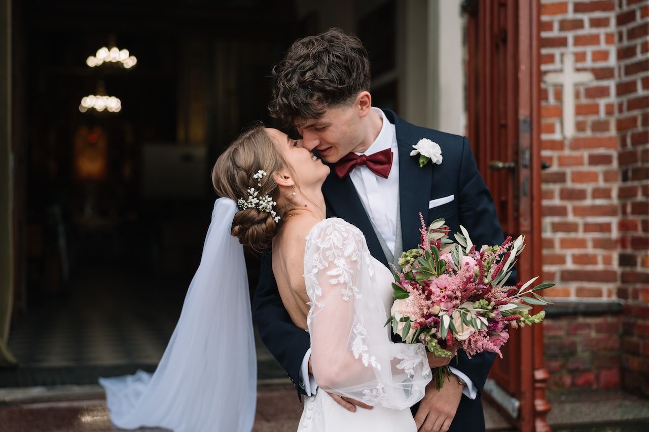 Newlyweds sharing a romantic kiss outside church entrance, bride holding colorful bouquet.