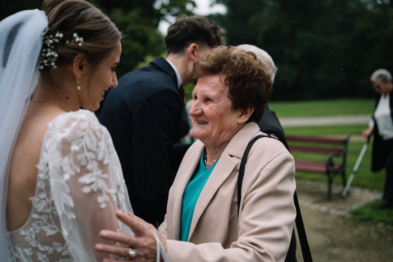 Elderly woman warmly greeting bride in white lace wedding dress at outdoor reception.