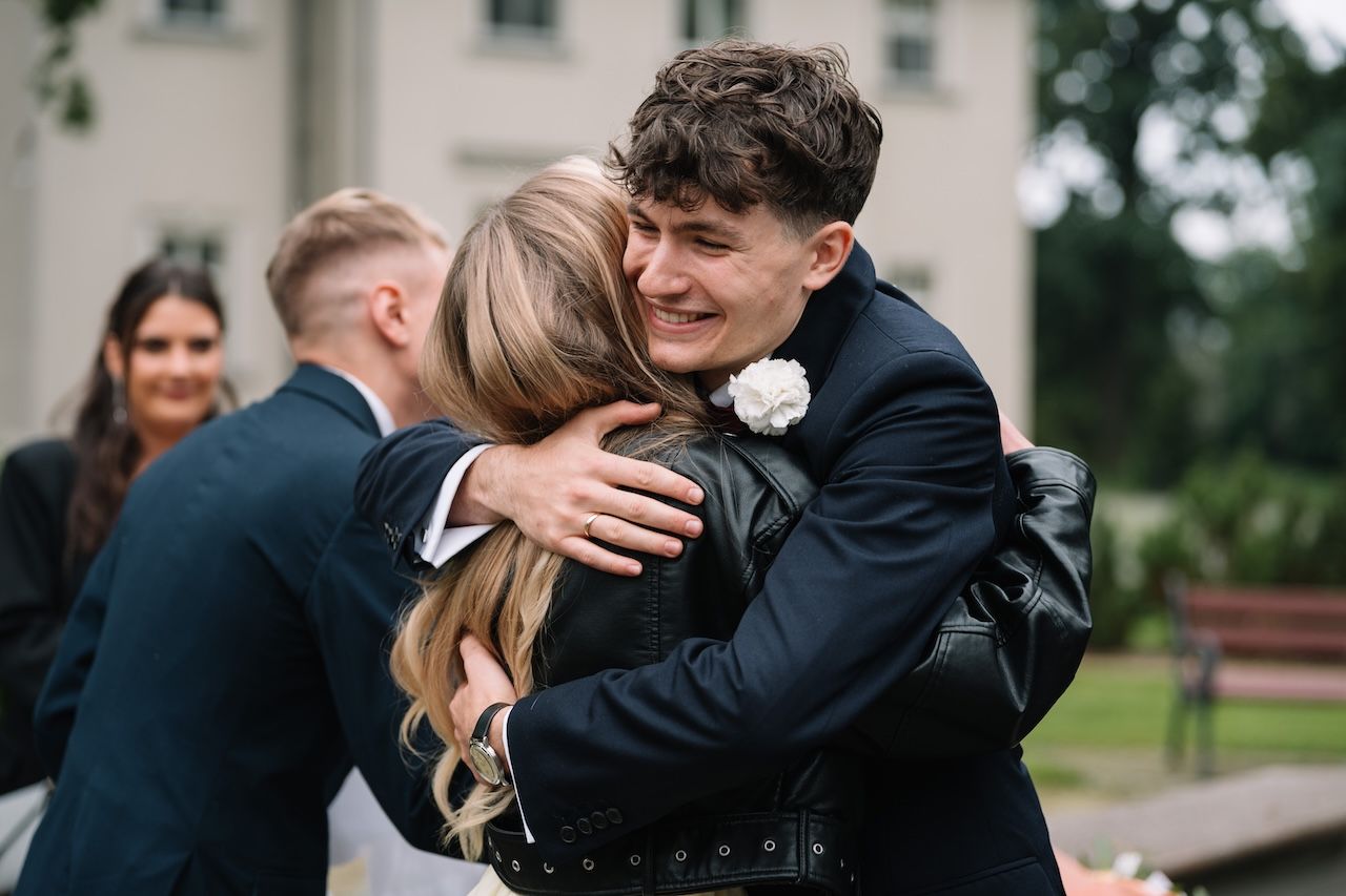 Groom in navy suit joyfully hugging wedding guest outdoors after ceremony celebration.