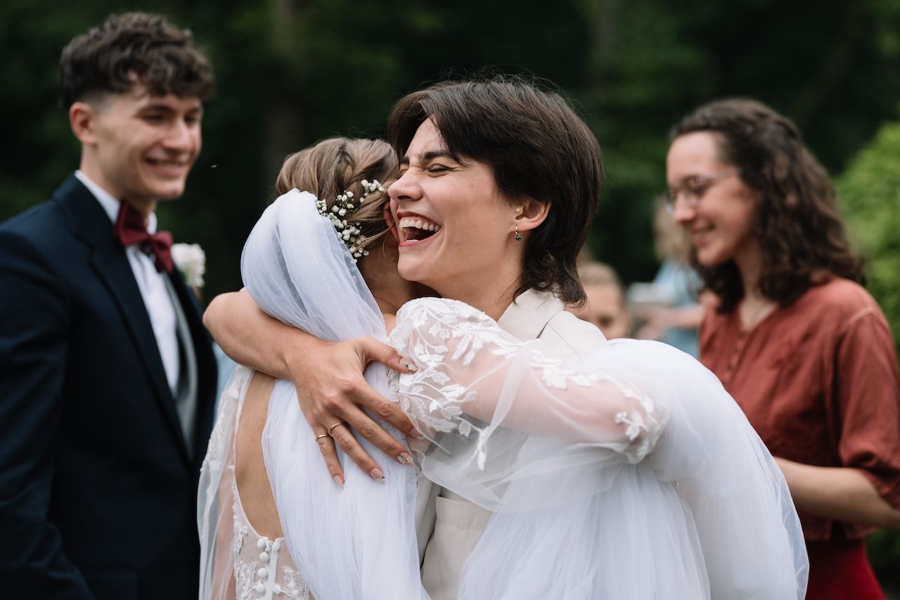 Bride in white lace gown laughing while embracing female wedding guest outdoors.