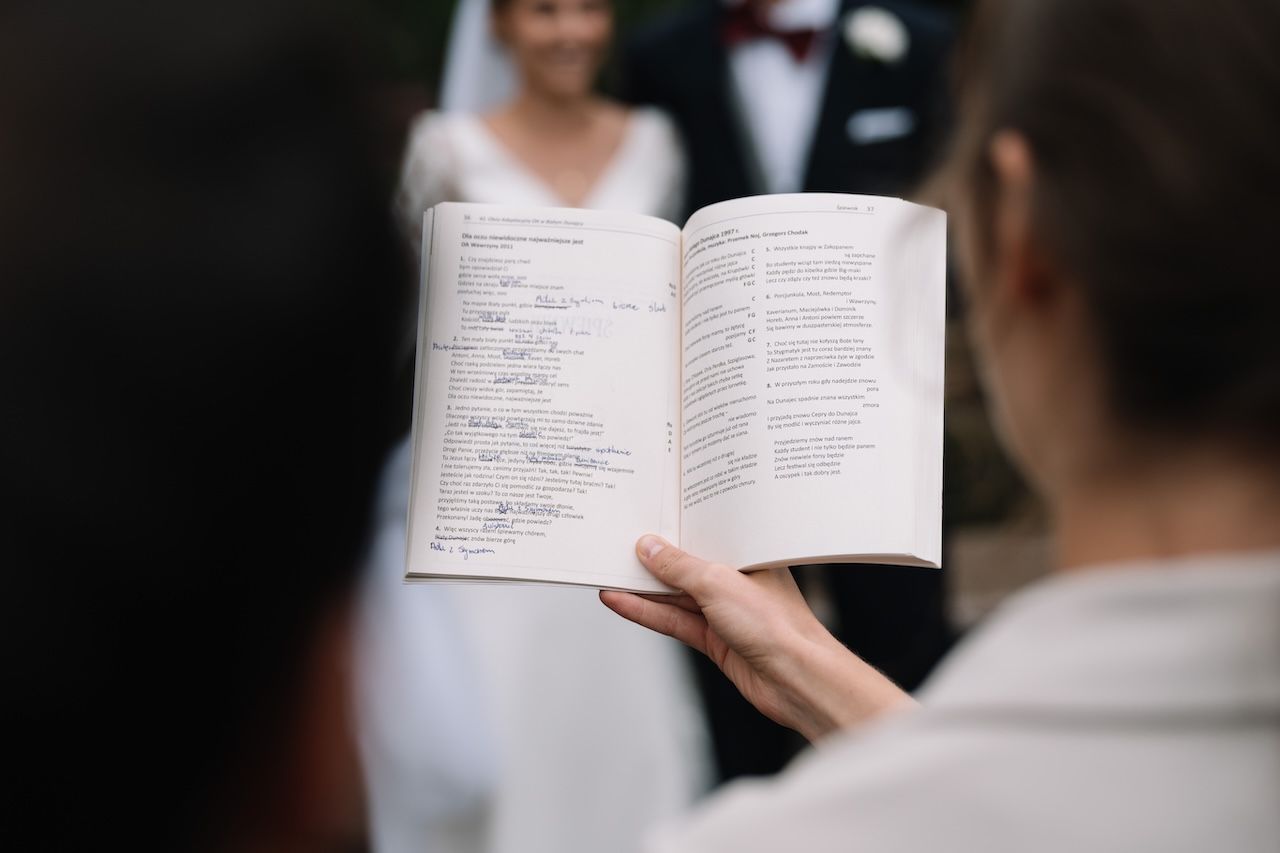 Officiant holding open ceremony book during outdoor wedding vows with couple in background.
