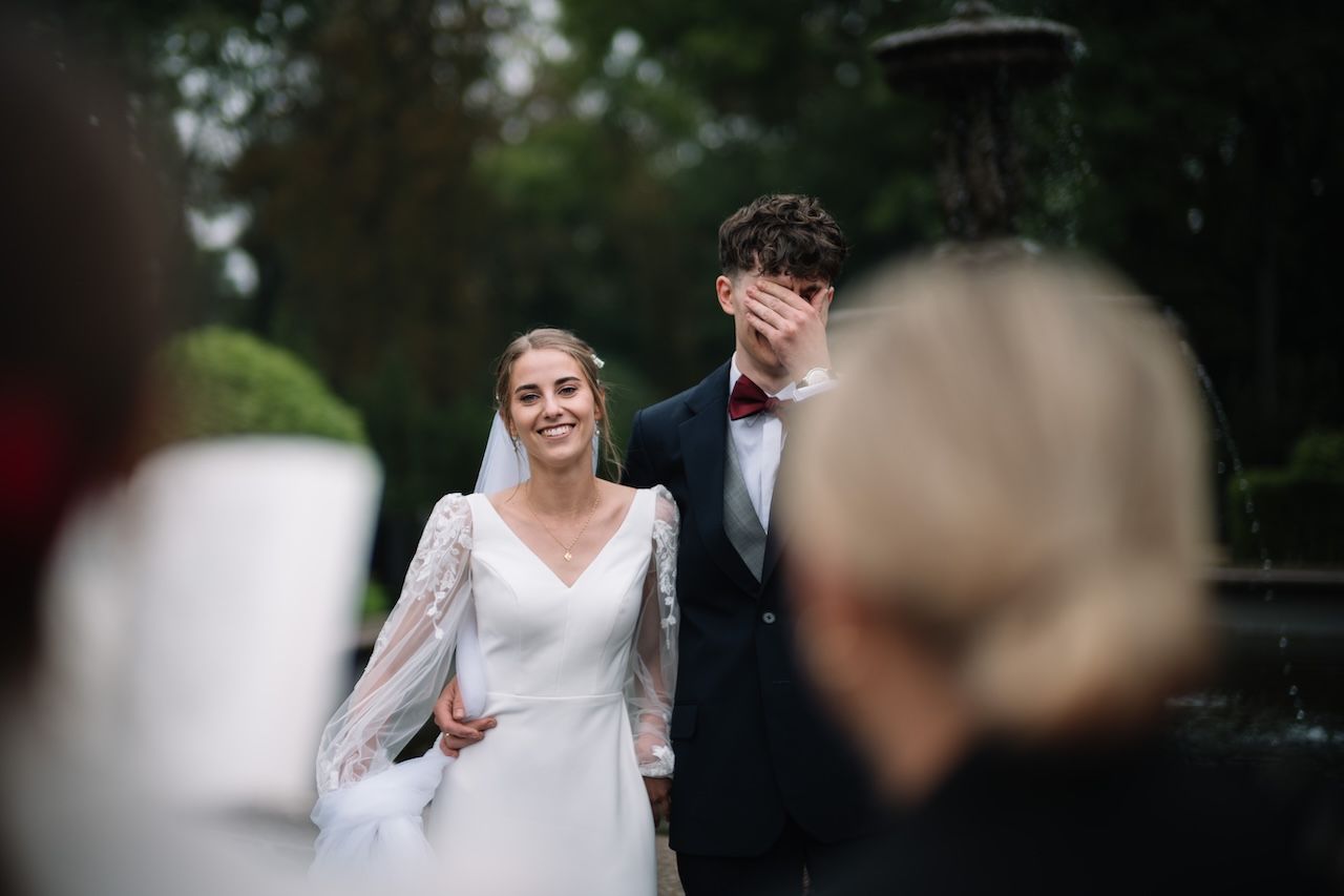 Smiling bride holding grooms hand outdoors as groom covers face near garden fountain.