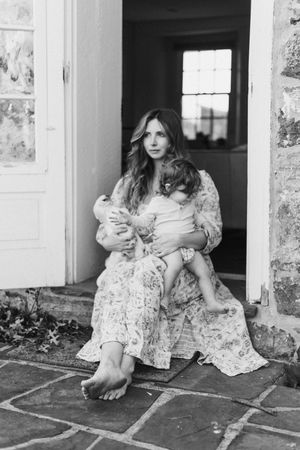 Black and white portrait of a woman in a floral dress sitting barefoot in a doorway with a baby.