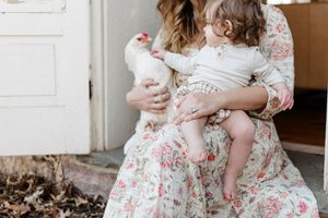 Parent in floral dress holding child and white chicken outdoors in natural light.