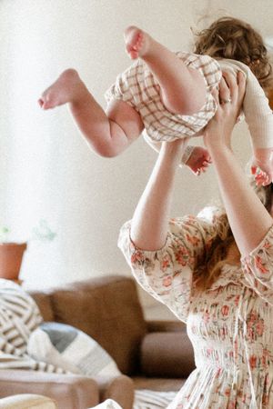 A baby in a checkered outfit is lifted playfully into the air while playing on a couch at home.