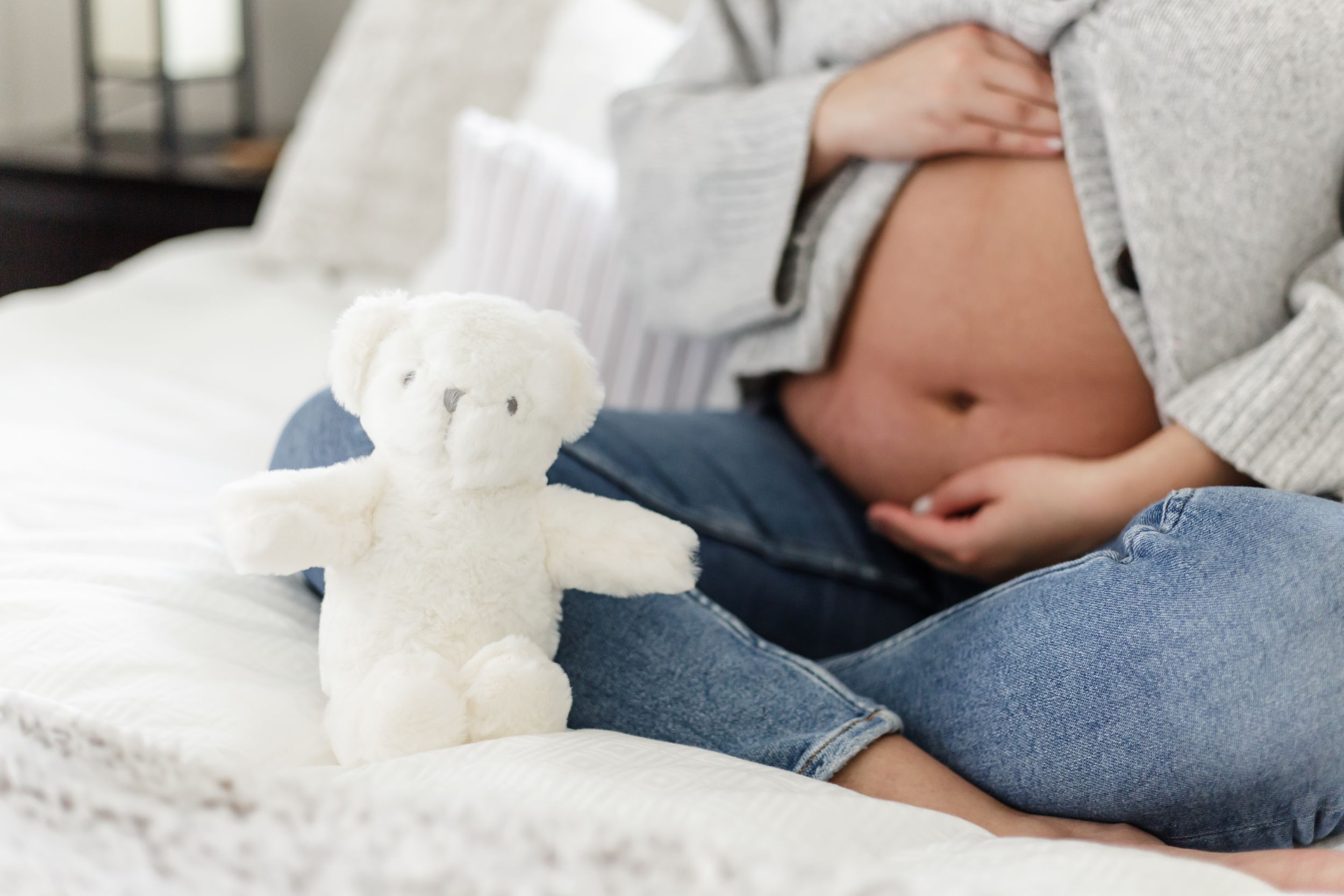 White teddy bear sits on bed while couple lounges in casual clothing against white bedding.