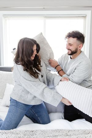 Couple in casual wear having fun with pillows on a bright white bed.