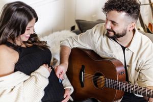 Person playing acoustic guitar while sitting on bed with companion in cozy setting.