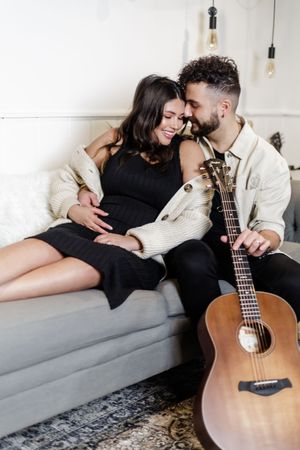 Couple lounging on gray couch with acoustic guitar in minimalist setting.