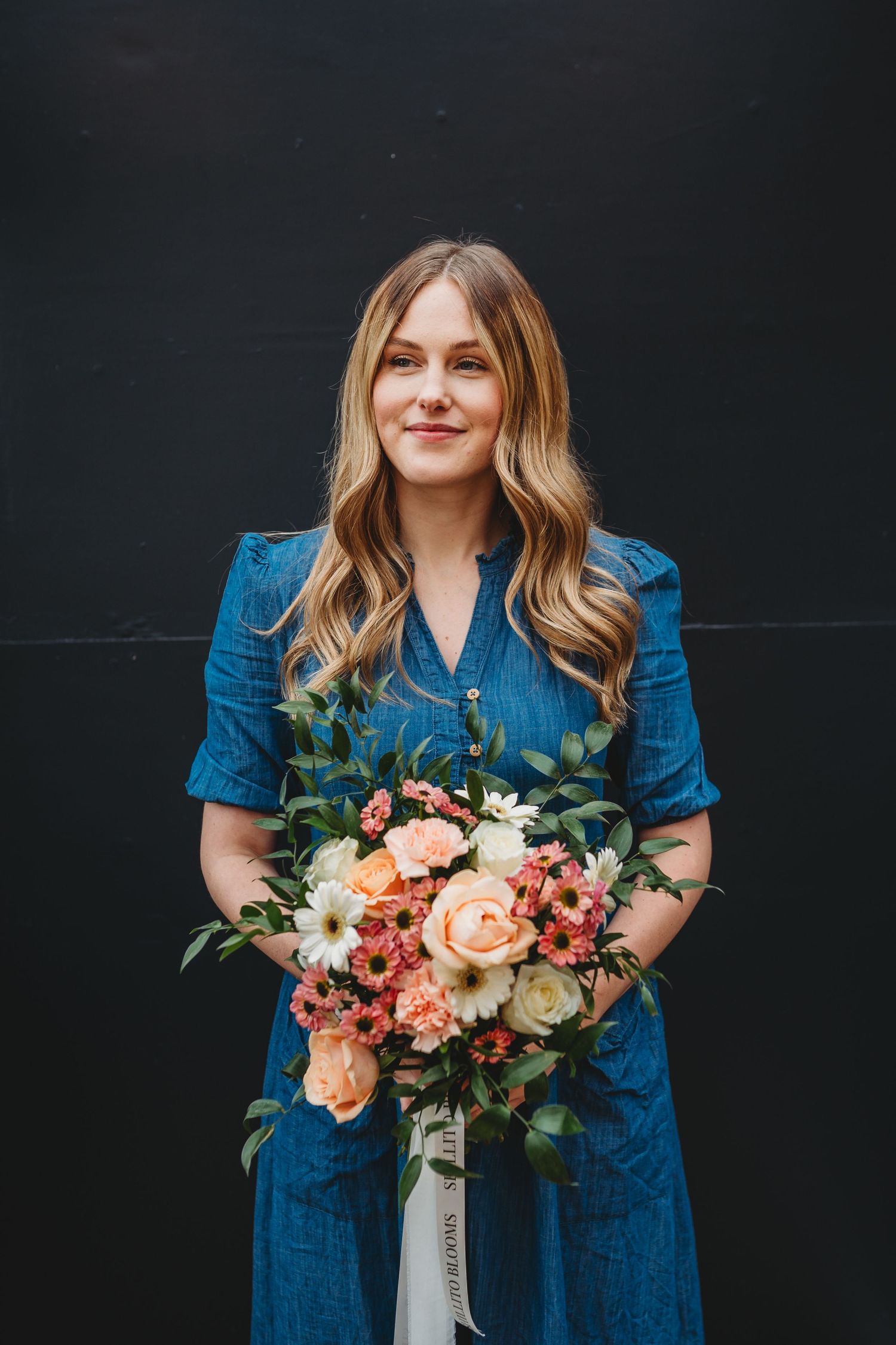 A person in a blue denim dress holds a vibrant bouquet of peach, coral and white flowers against a dark background.