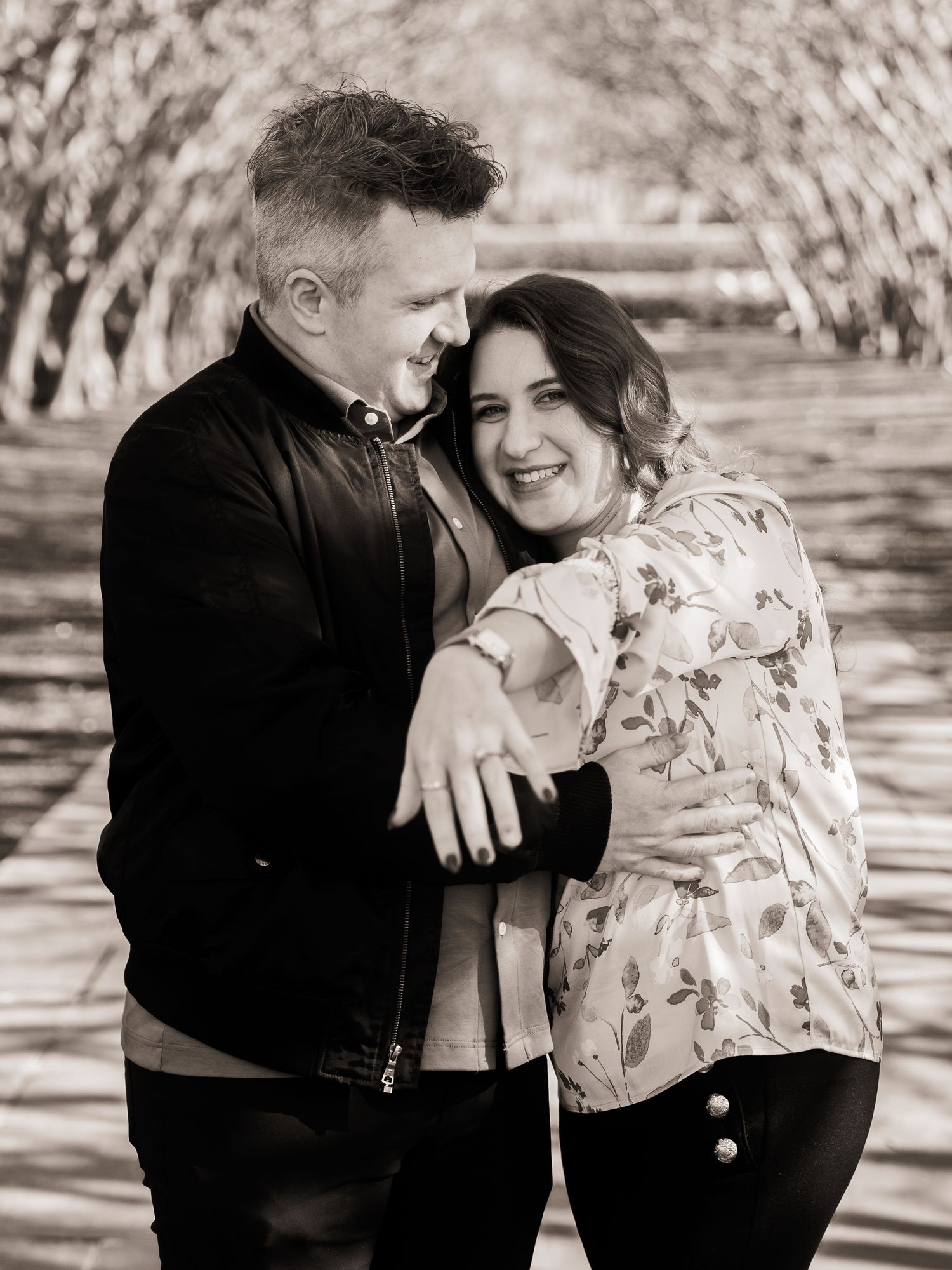 Happy engaged couple in black and white photo sharing romantic embrace under arched tree branches.