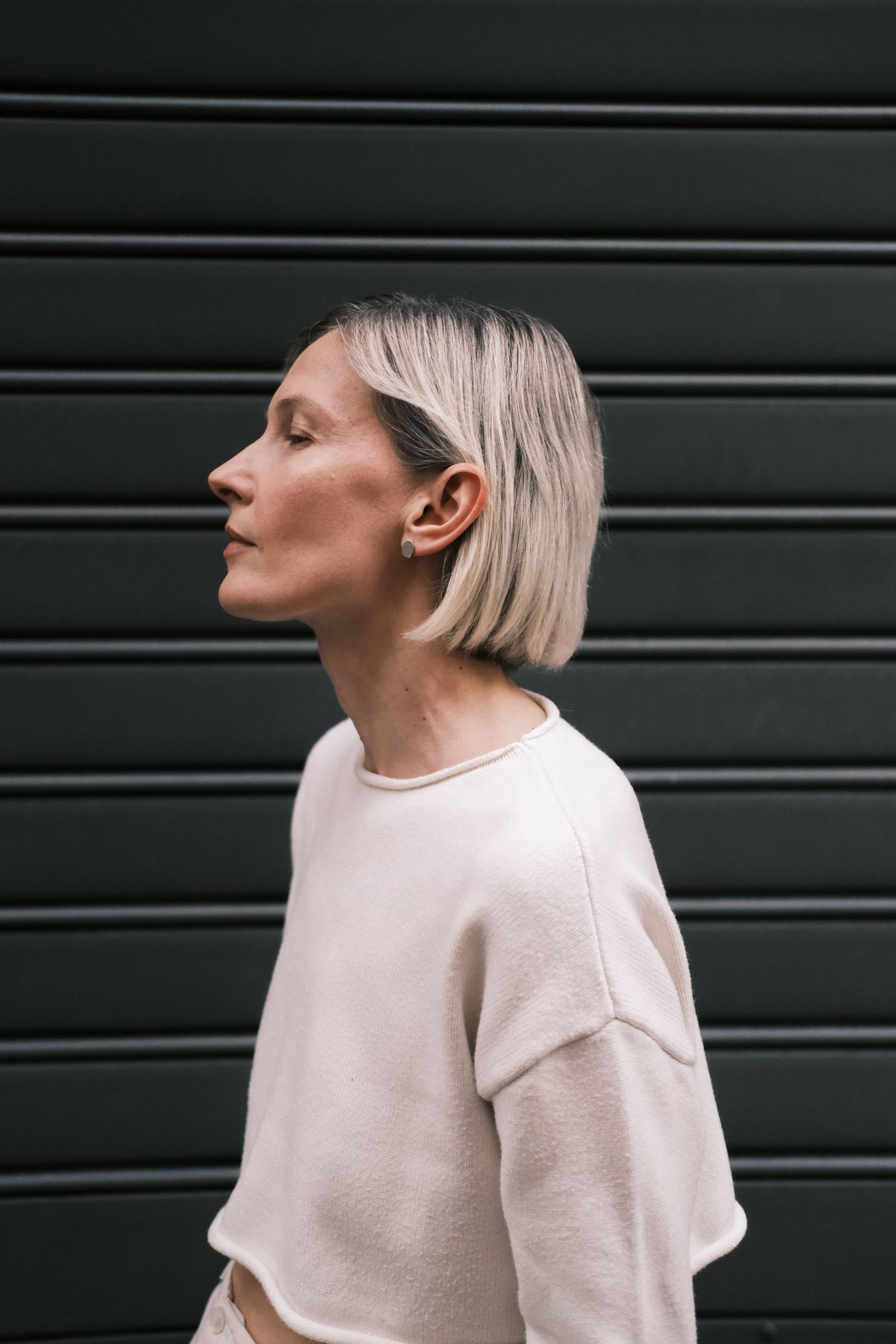 Side profile of person with platinum blonde bob haircut against dark green industrial background.