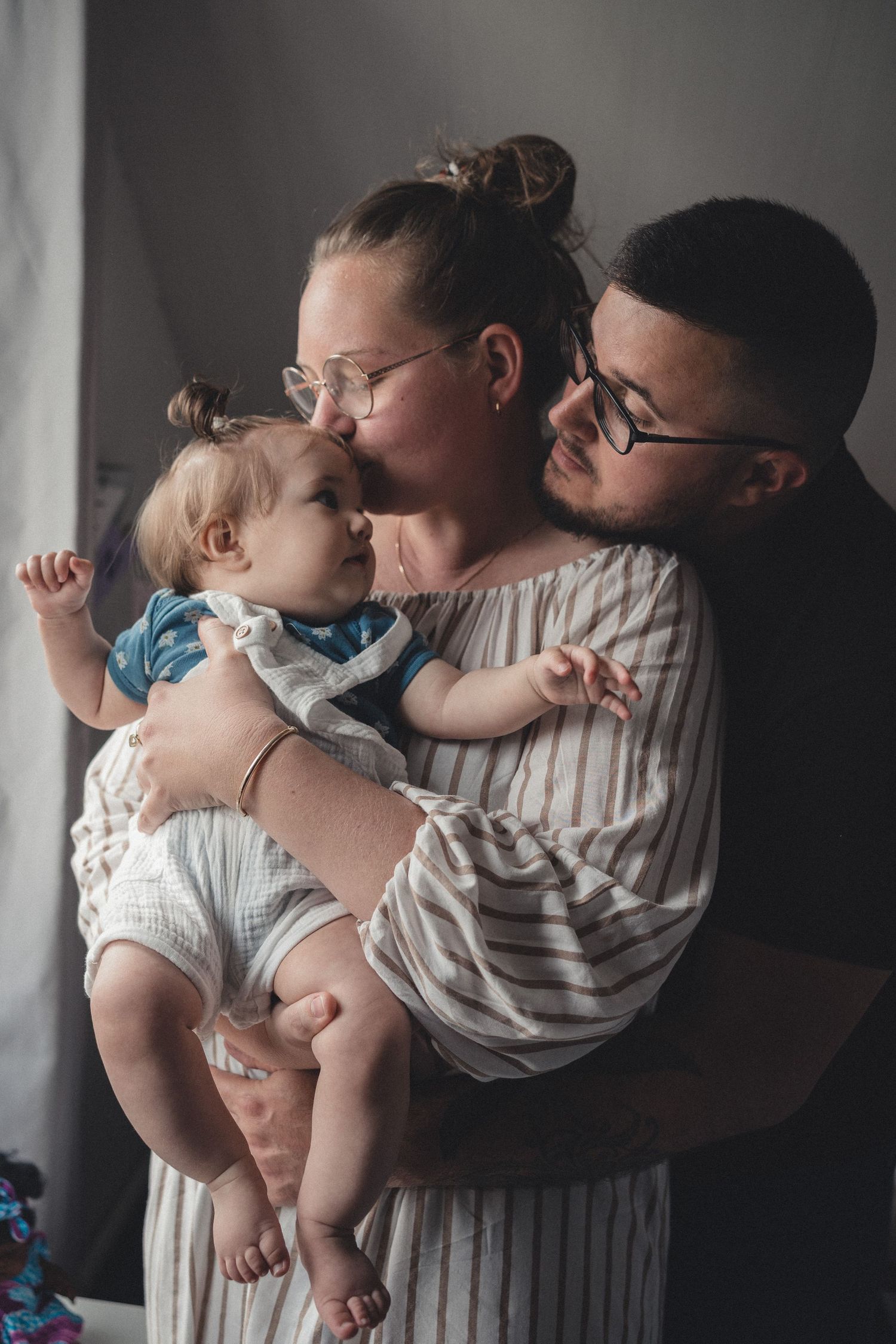 A tender family moment captured in soft lighting as parents hold and kiss their baby in matching striped outfits.
