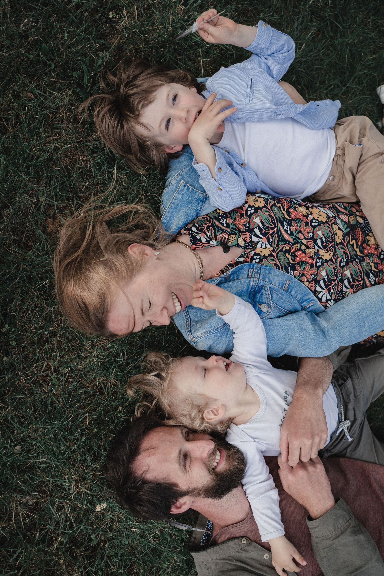 Moment en famille avec les parents et leurs enfants, tous allongés dans l'herbe les uns à côté des autres. Ils rient et se regardent.