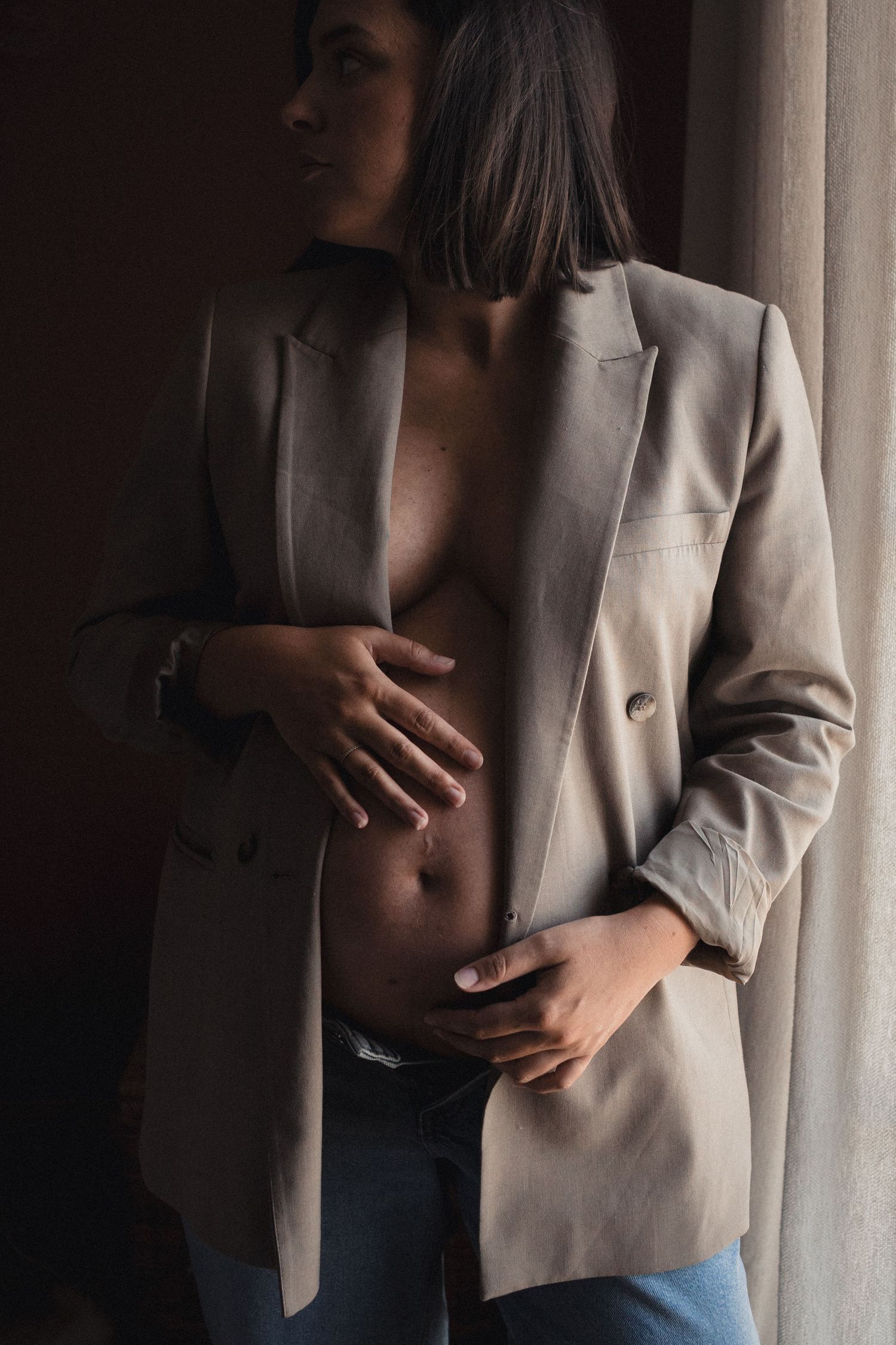 A person in a beige blazer and jeans stands against a light curtain background showing their midsection.
