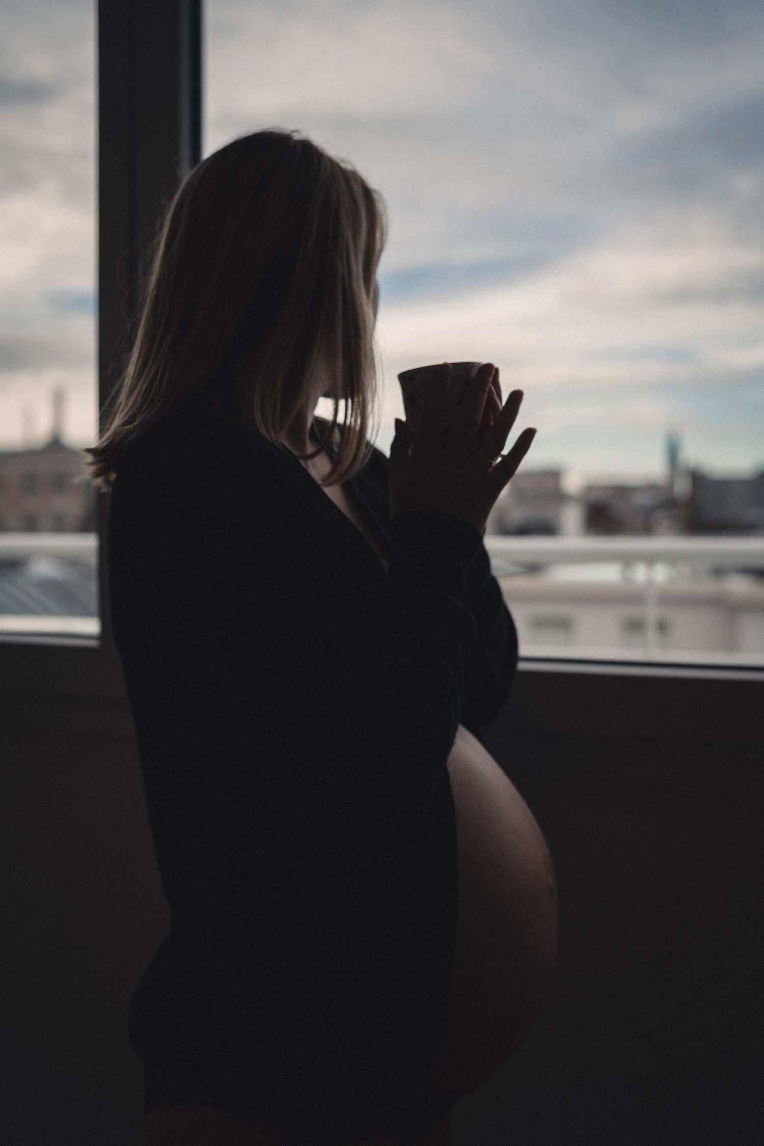 1 femme enceinte regarde les toits de la ville par la fenêtre en tenant une tasse de café en contre jour. Gilet noir et ventre rond.
