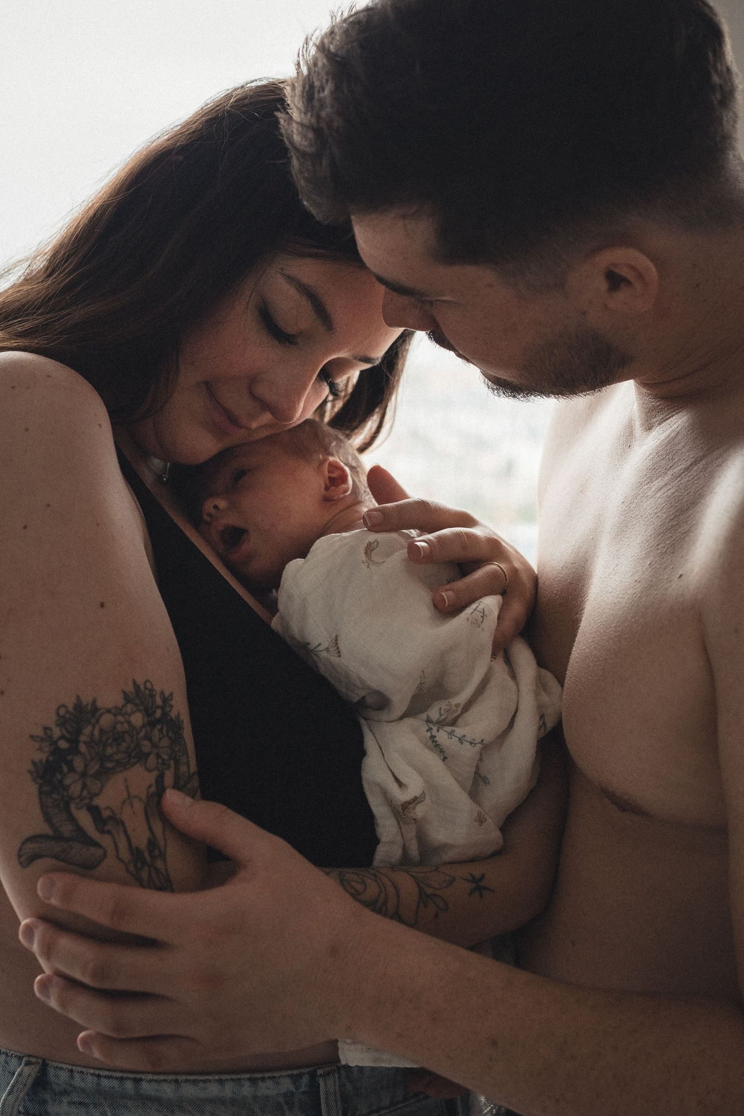 Une famille en contre jour: la maman pose sa joue sur la tête de son bébé nouveau-né et le papa les entoure de son bras en les regardant