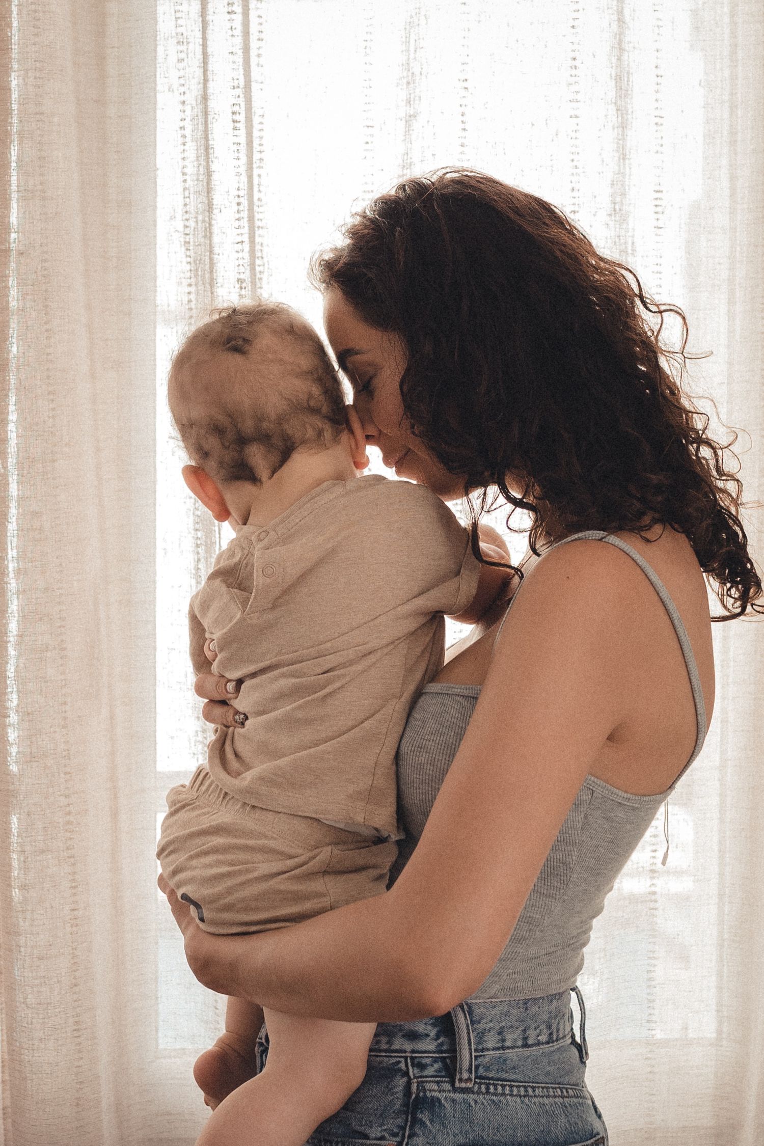 A tender moment between parent and child silhouetted against sunlit curtains in a warm, intimate indoor setting.