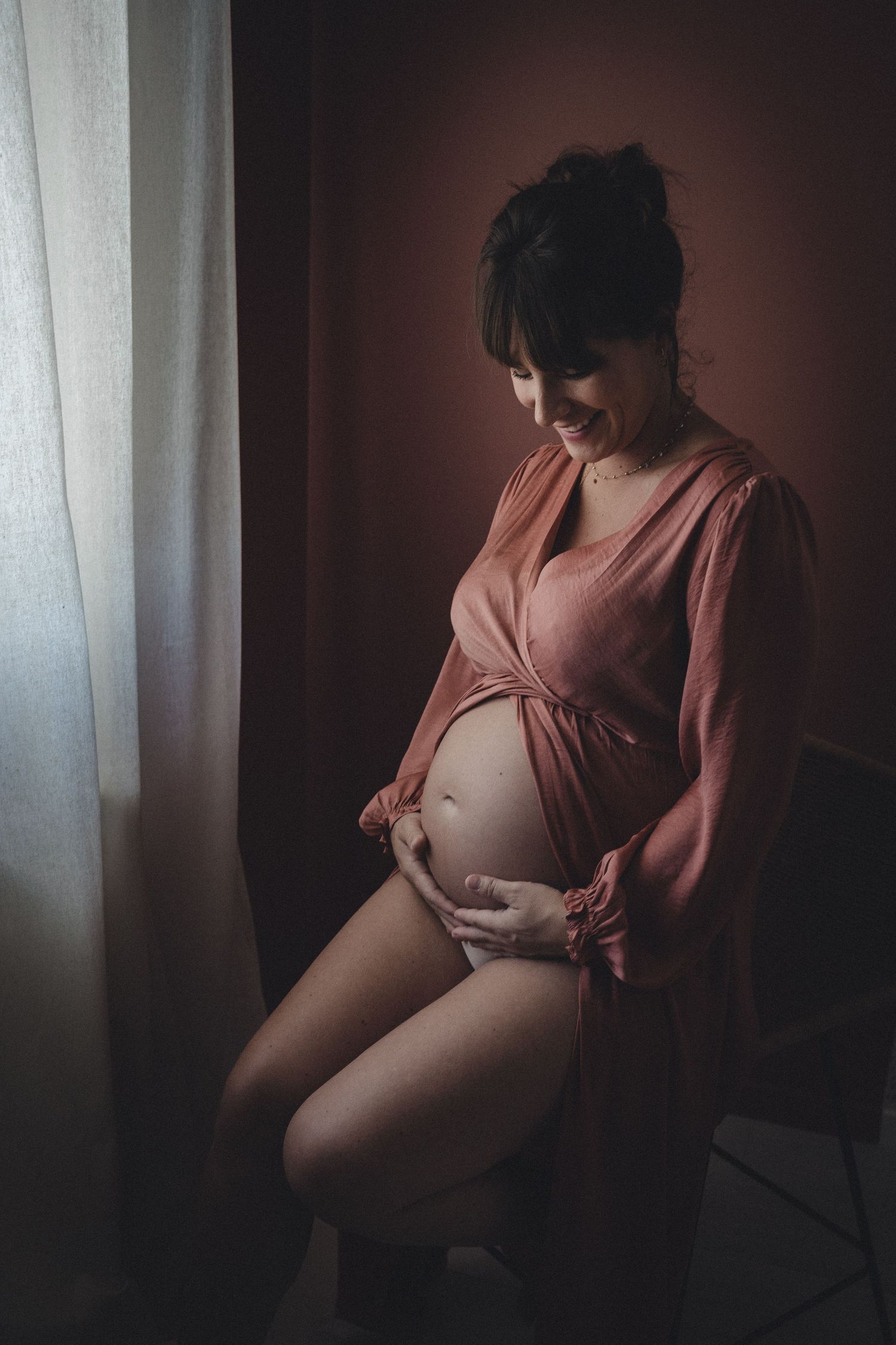 1 femme enceinte dans une robe en soie rose foncé regarde son ventre nu et sourit près d'une fenêtre à la lumière diffuse