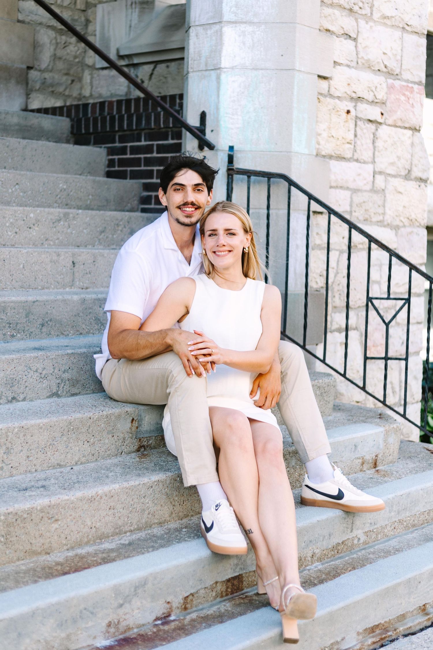 Young couple in casual white outfits sitting together on stone steps of a building.