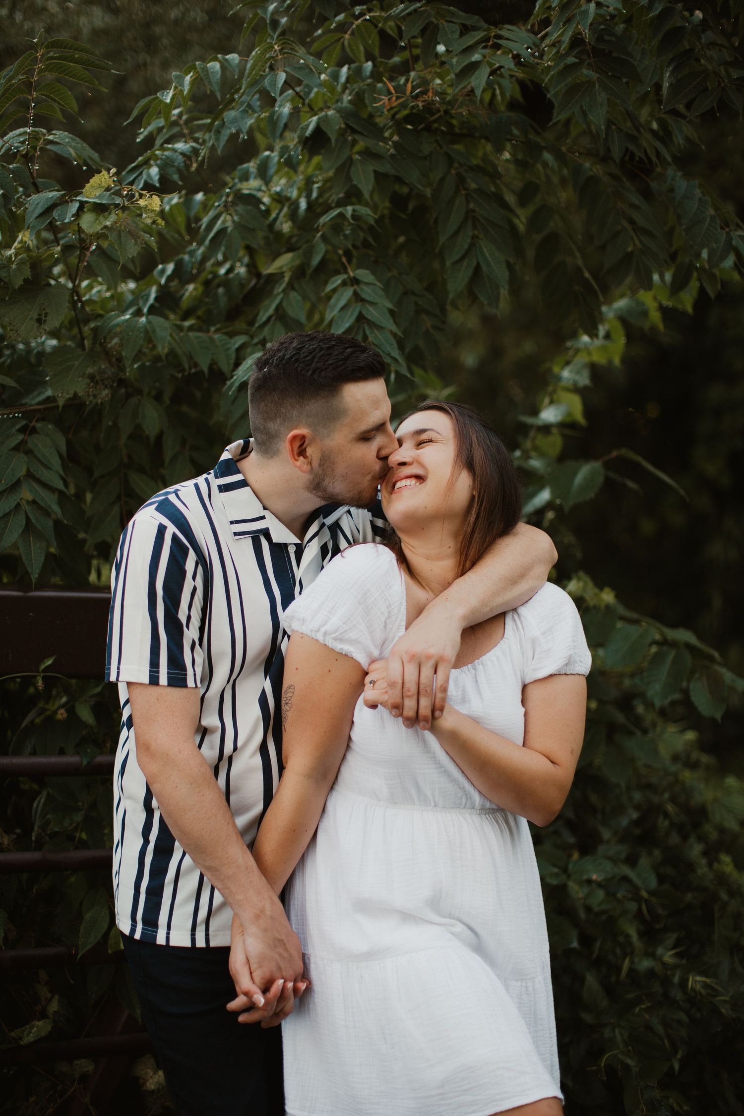 A couple shares an intimate moment outdoors, with the person in stripes embracing their partner in white against green foliage.