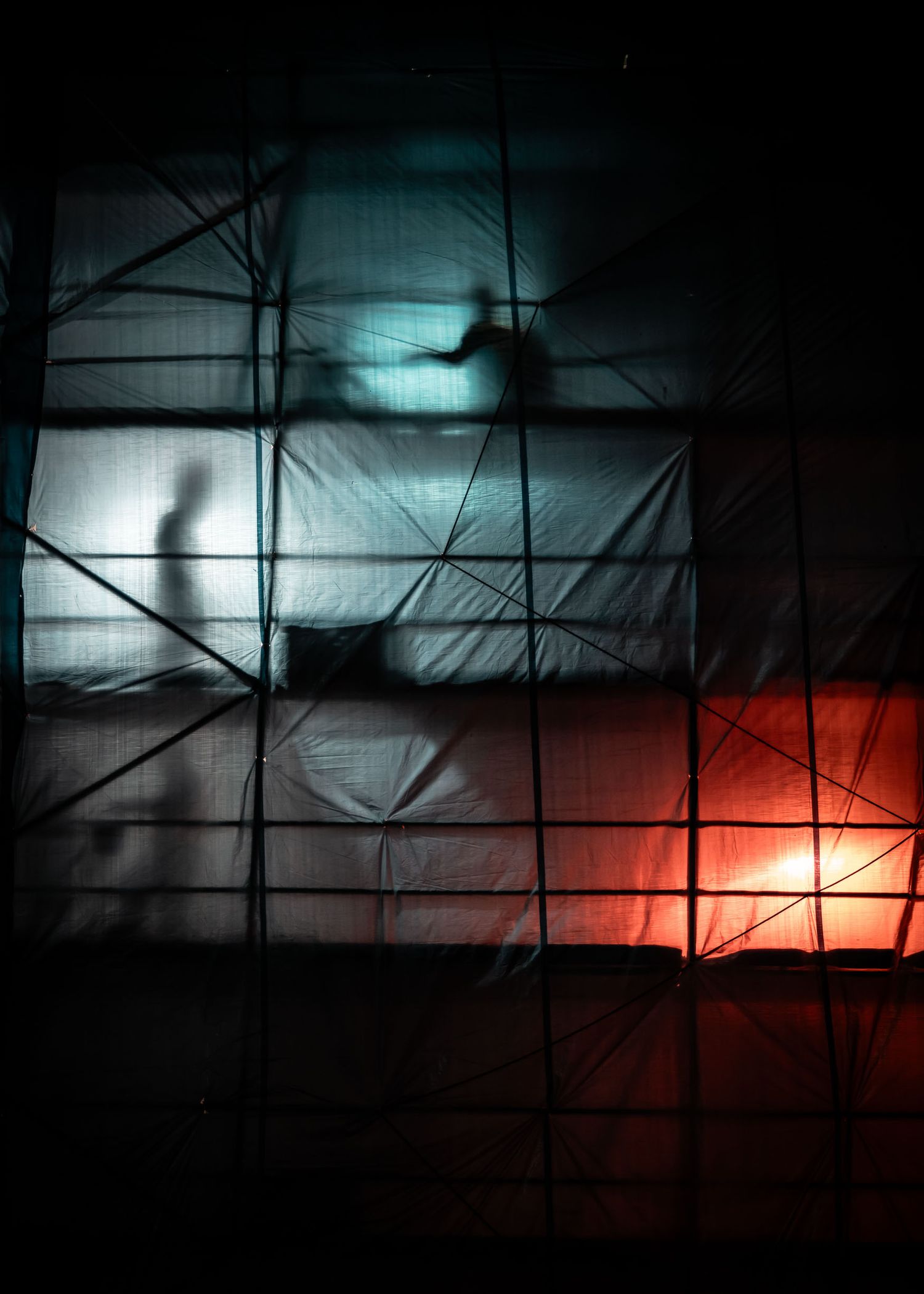 Dark silhouettes cast dramatic shadows against an illuminated backdrop of teal and red light through translucent fabric panels.