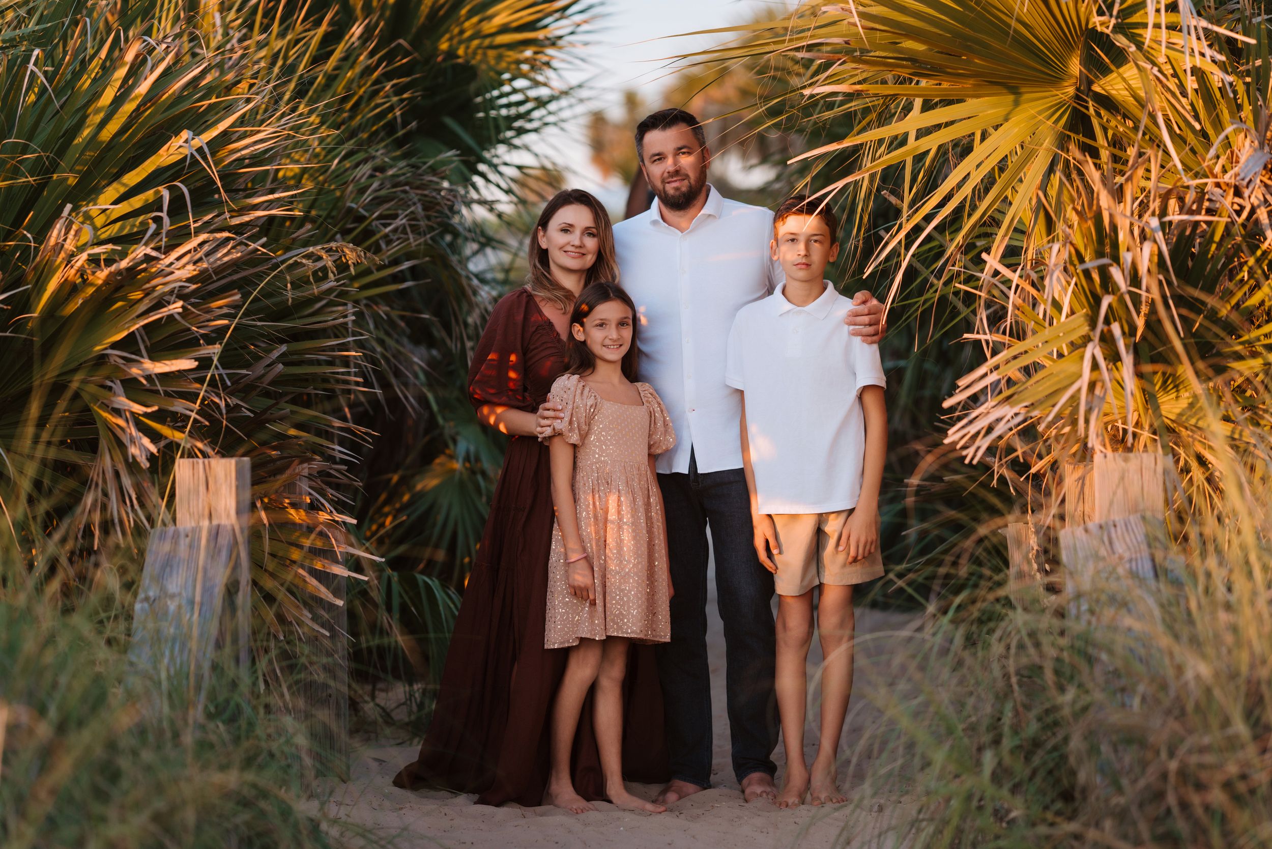 A family group poses together on a path surrounded by palm trees at sunset with warm golden lighting.