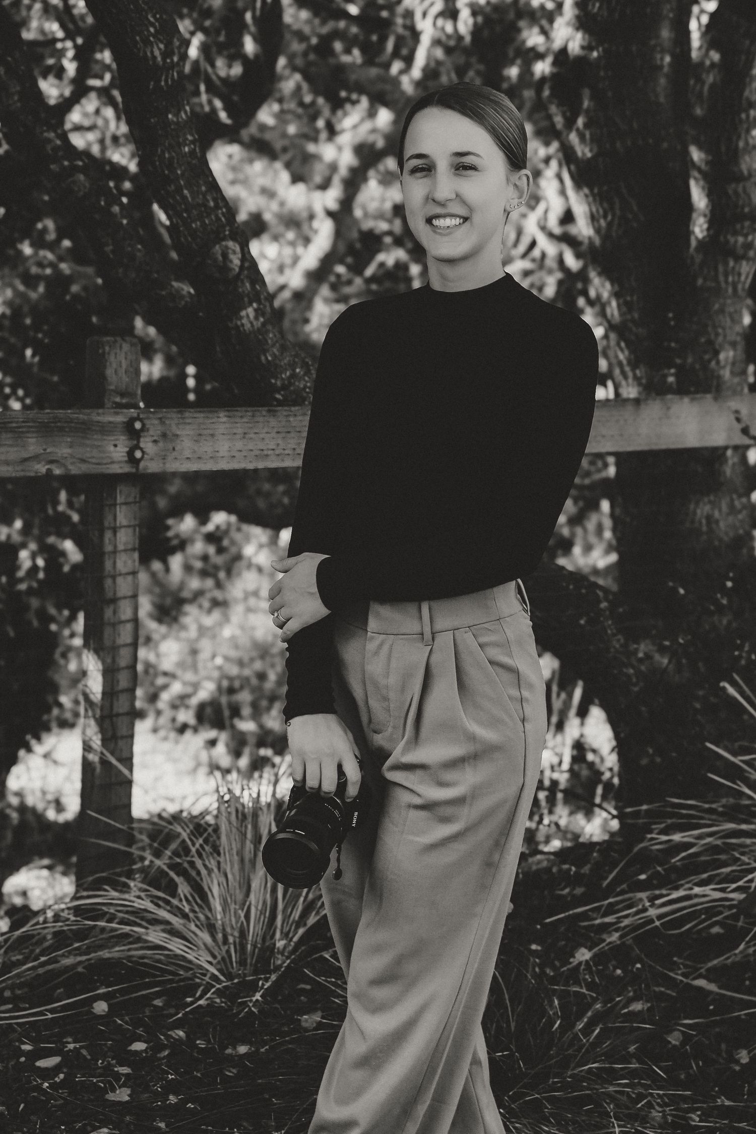 Black and white portrait in outdoor setting showing person in black turtleneck and khaki trousers holding camera.