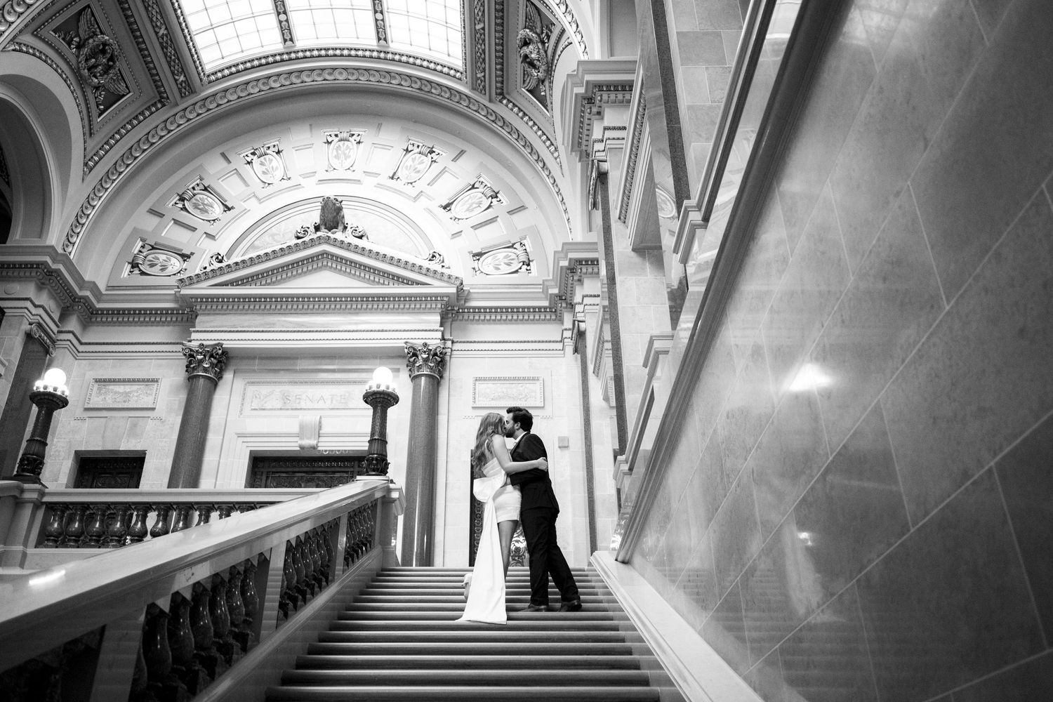 A romantic black and white photo of two people embracing on an elegant marble staircase beneath ornate classical architecture.