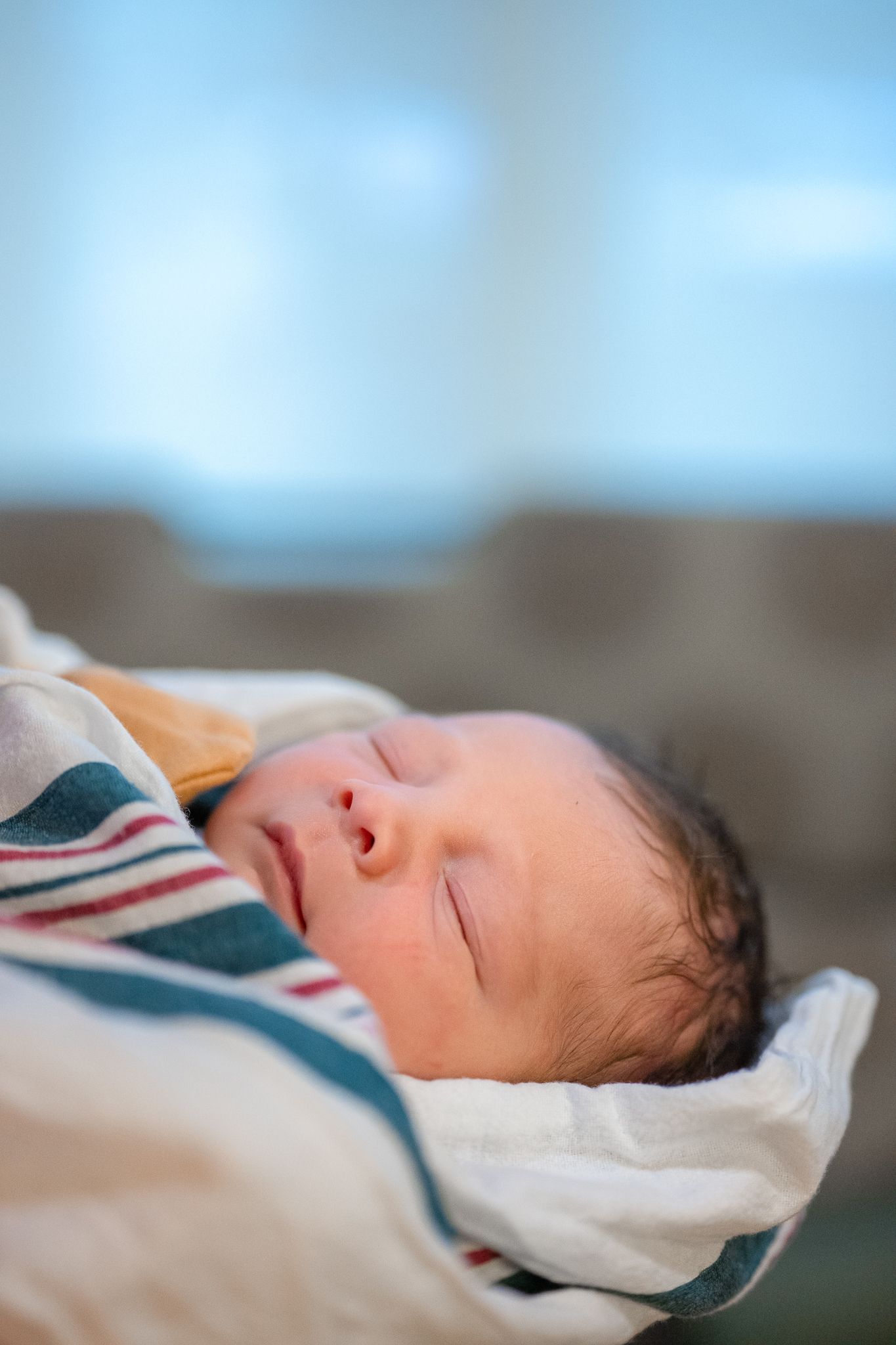 A sleeping newborn baby wrapped in a white and striped blanket rests peacefully against a soft background.