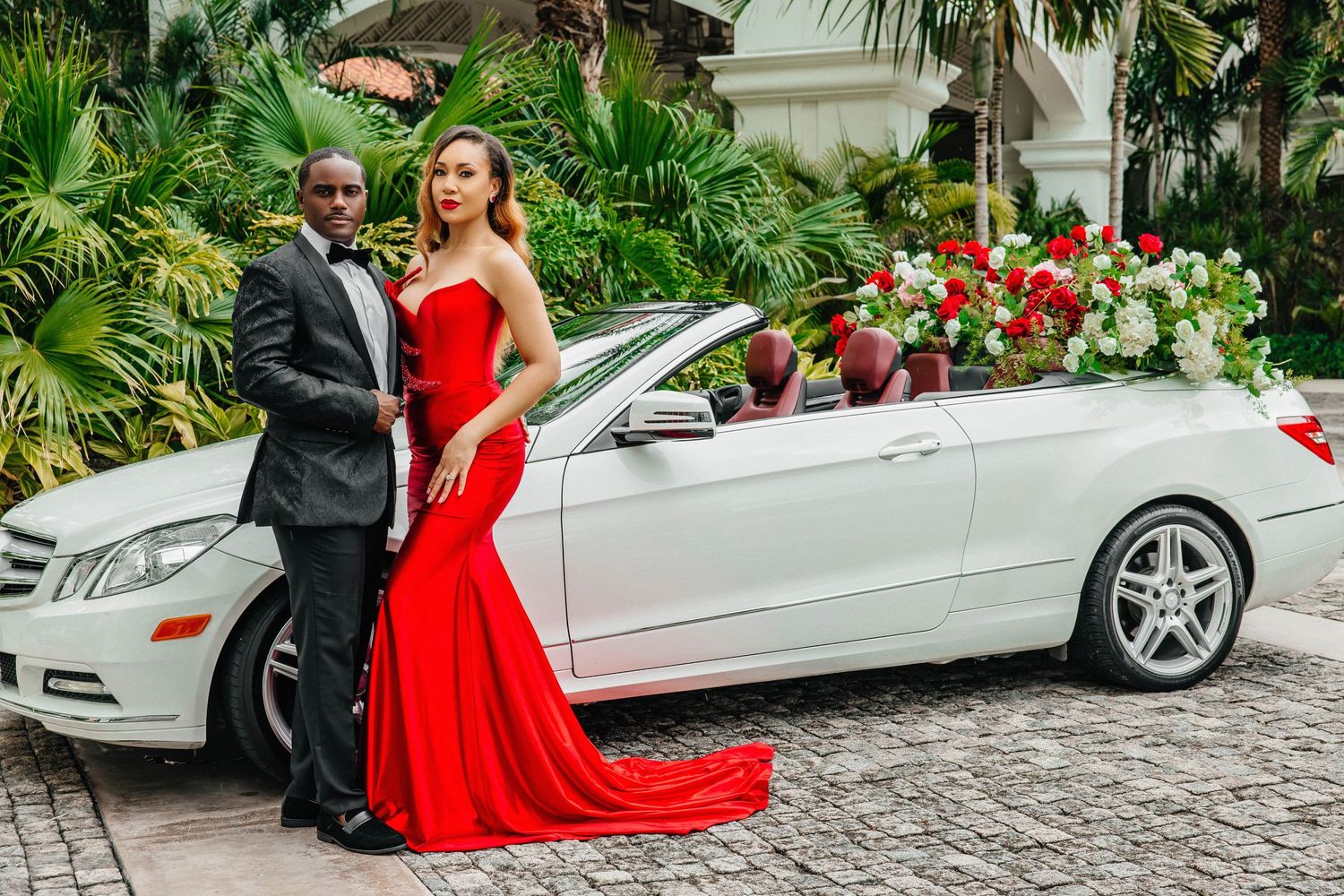 Couple poses with decorated white Mercedes, featuring dramatic red gown with long train.