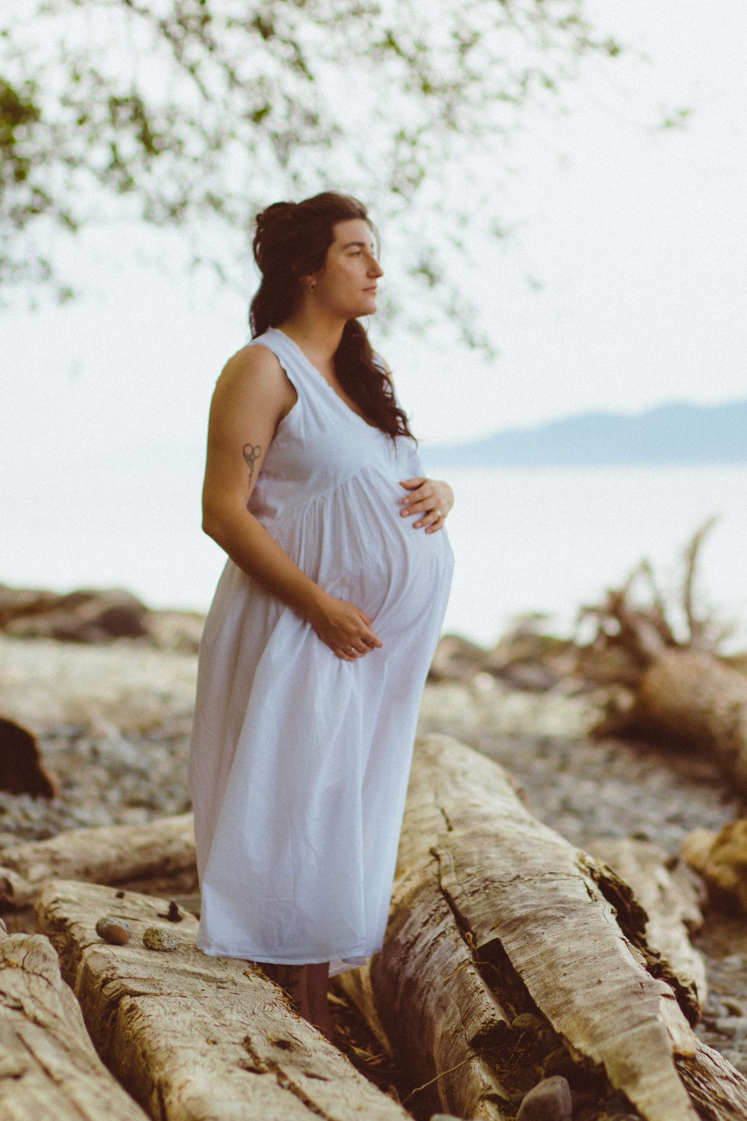 Pregnant mom in white flowing dress touching her belly while overlooking the ocean in Vancouver, BC during an outdoor maternity photoshoot.