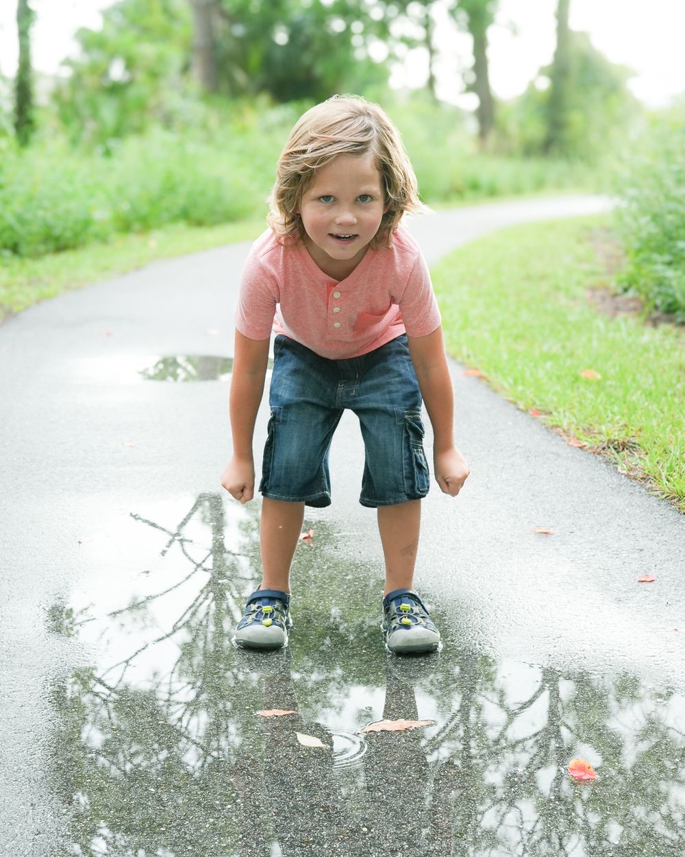 A playful child crouches over a puddle on a wet path surrounded by greenery.