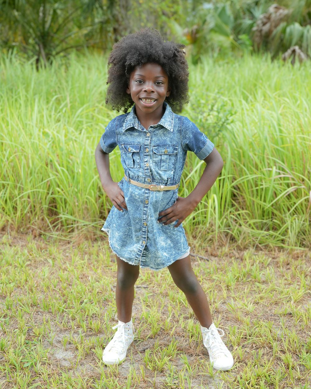 Child in denim romper strikes confident pose in grassy field while wearing white sneakers.