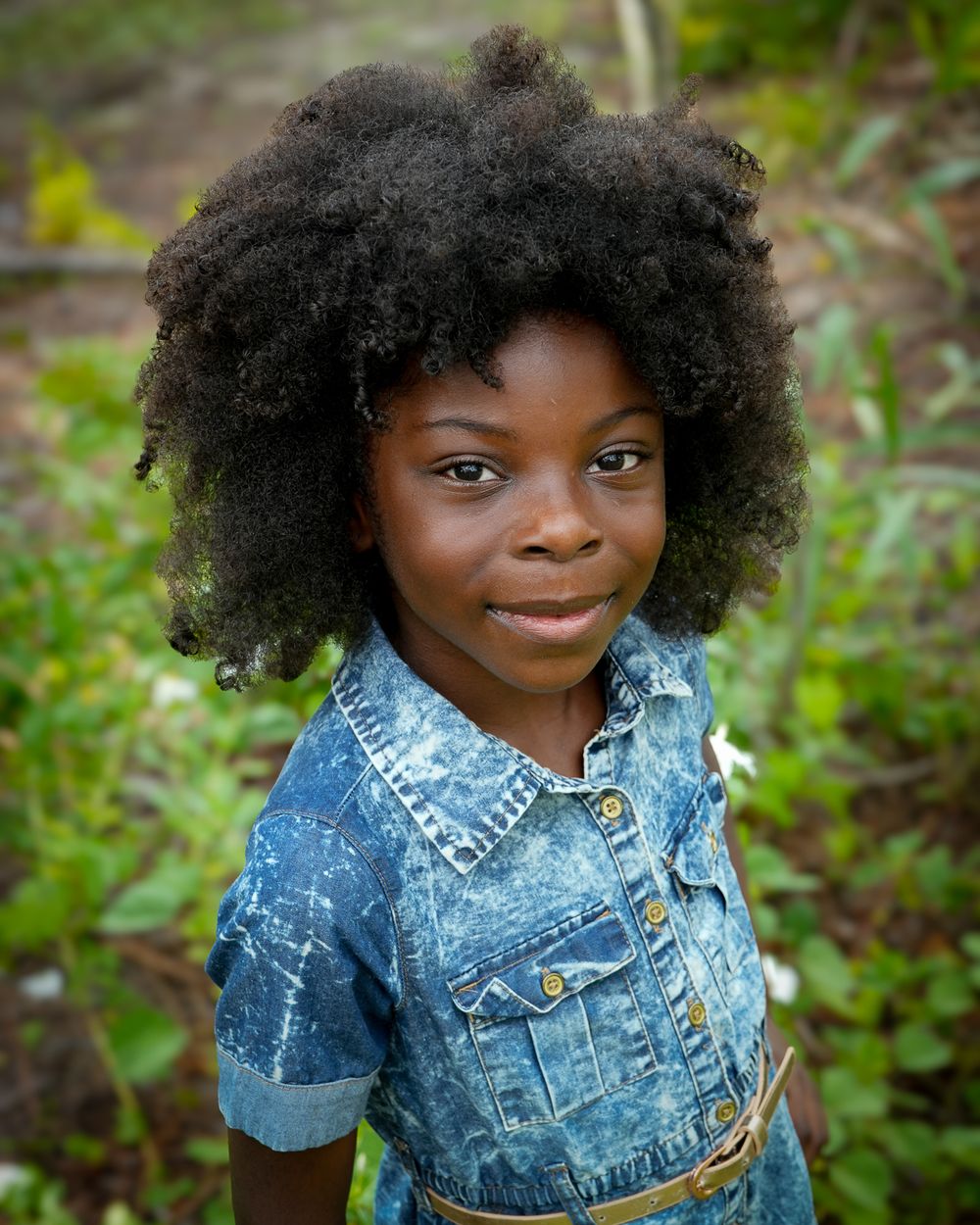 A young person with natural curly hair poses in a denim jacket against a green background.