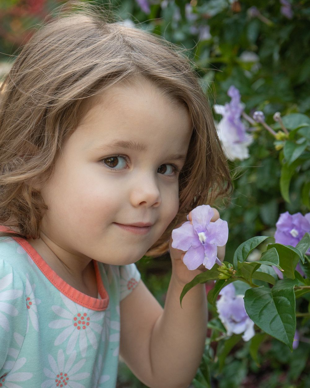 Young child admiring purple flowers in a garden while wearing a mint floral dress.