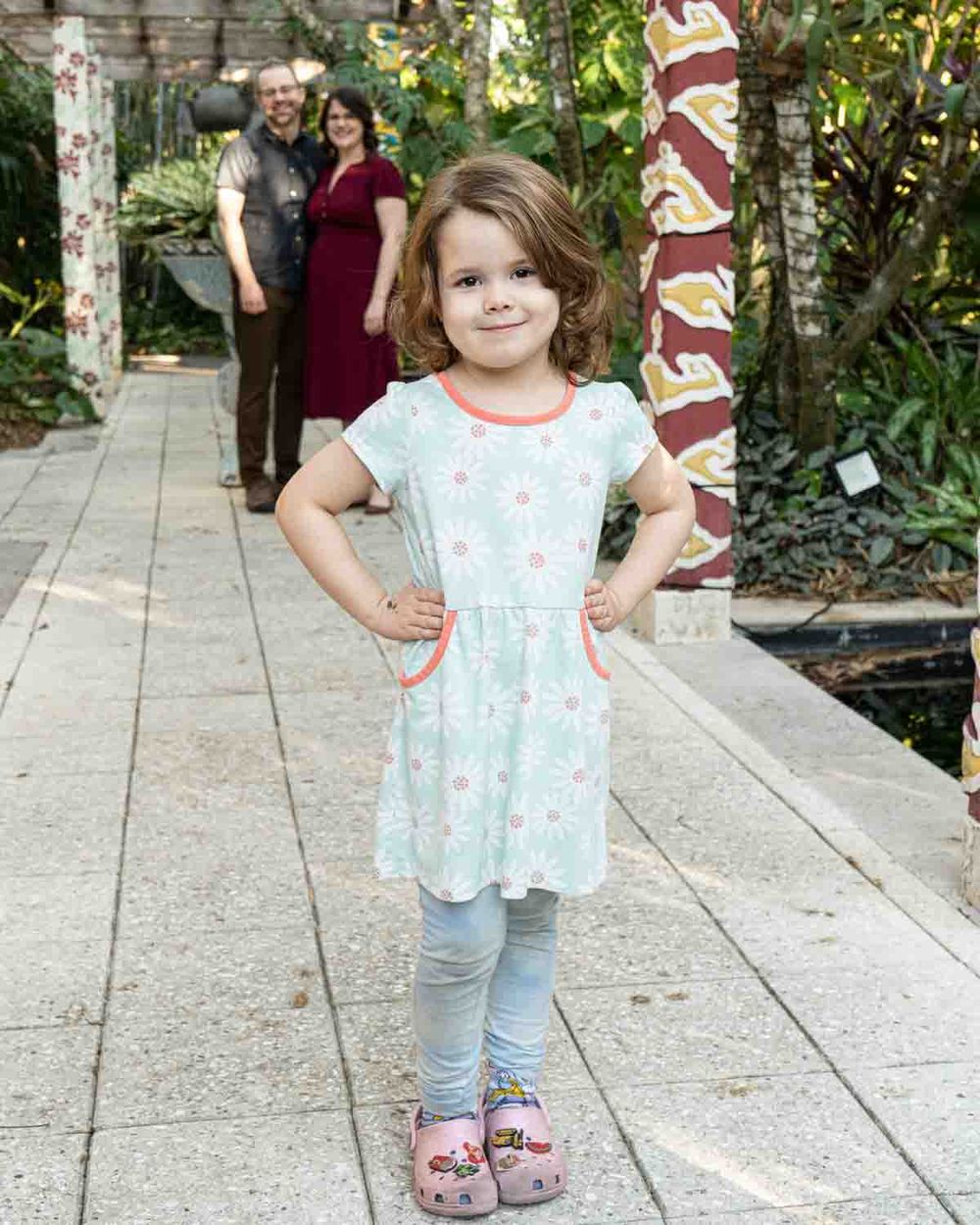 A young child poses confidently in a floral dress and leggings on a garden path.