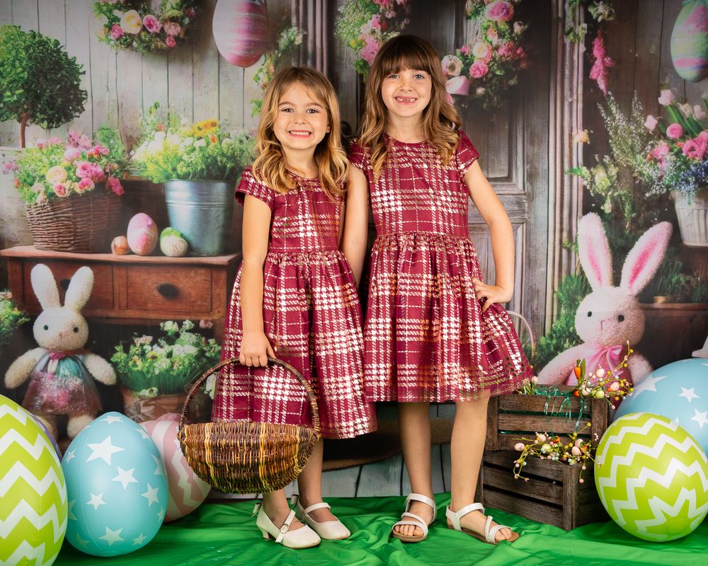 Two children in matching red plaid dresses pose with Easter decorations and colorful eggs.