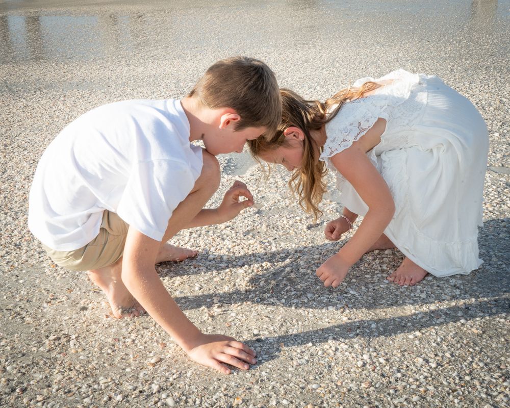 Two children in white outfits bend down to examine something on a sandy beach.