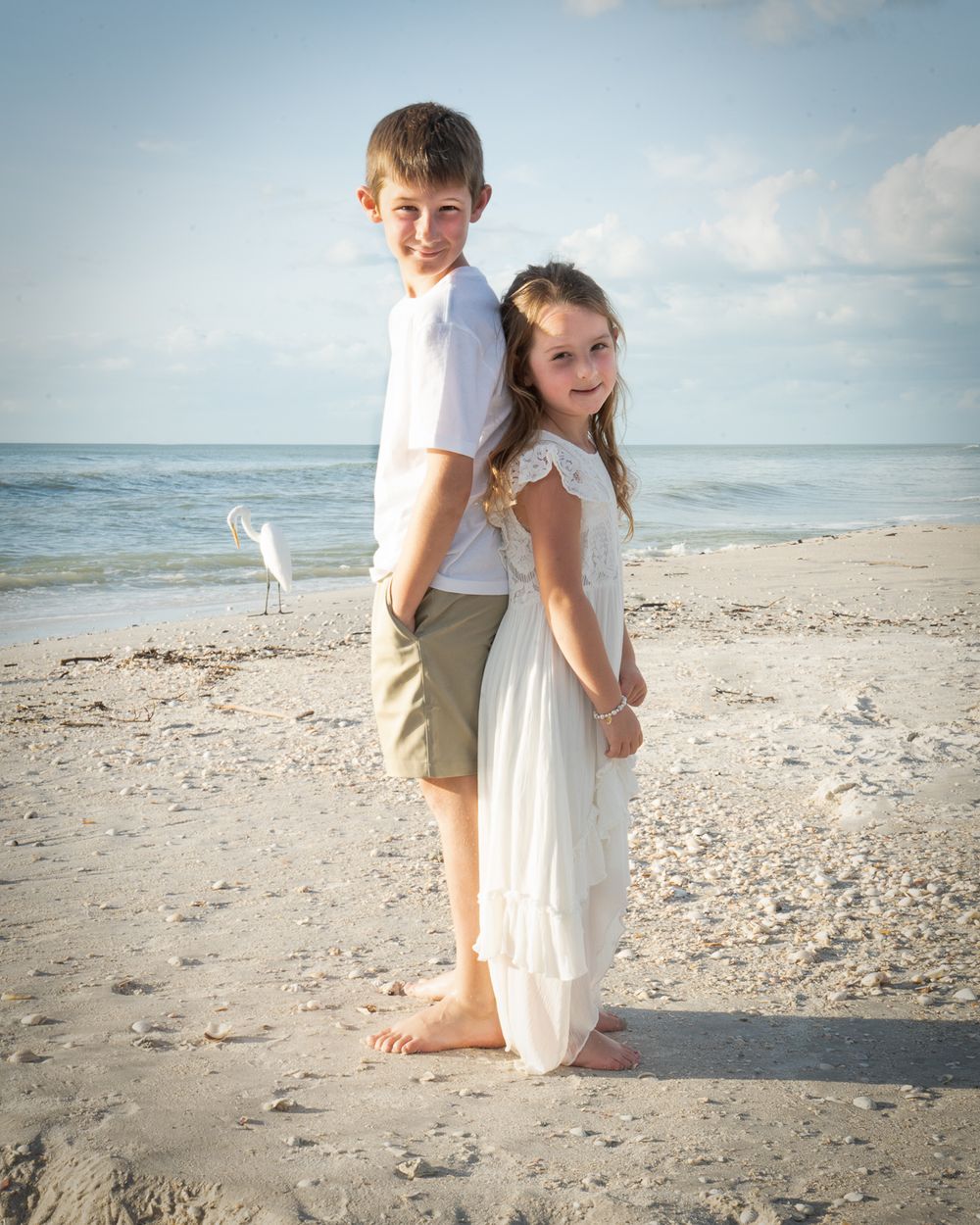 Two children in casual white outfits pose playfully back-to-back on a beach with ocean view.