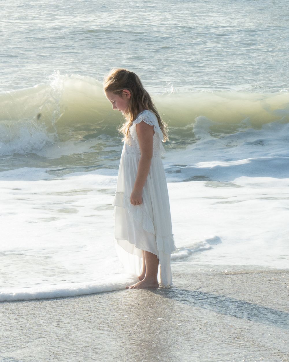 A girl in a white dress stands at the ocean's edge as waves roll in.