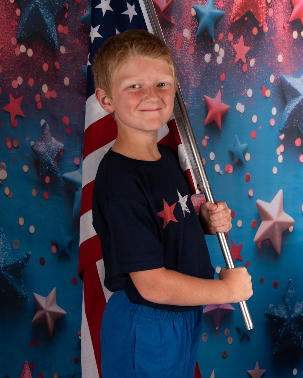 Child posing with American flag against star-spangled backdrop in navy outfit.