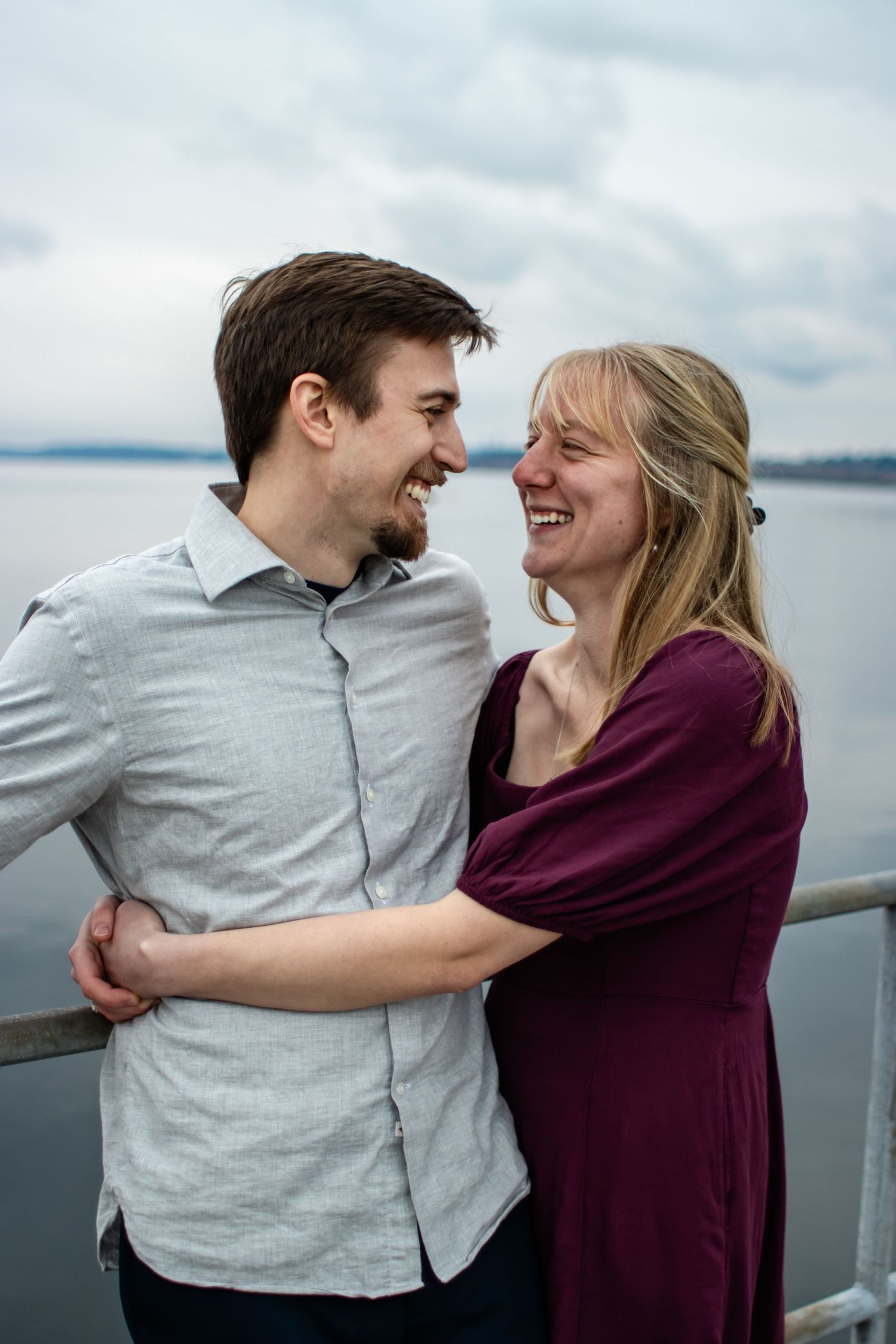 Laughing couple embrace on waterfront dock with cloudy sky and lake in background.