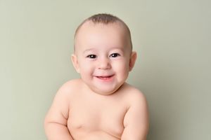 Adorable baby smiling while posing shirtless against light green background during professional photo session.