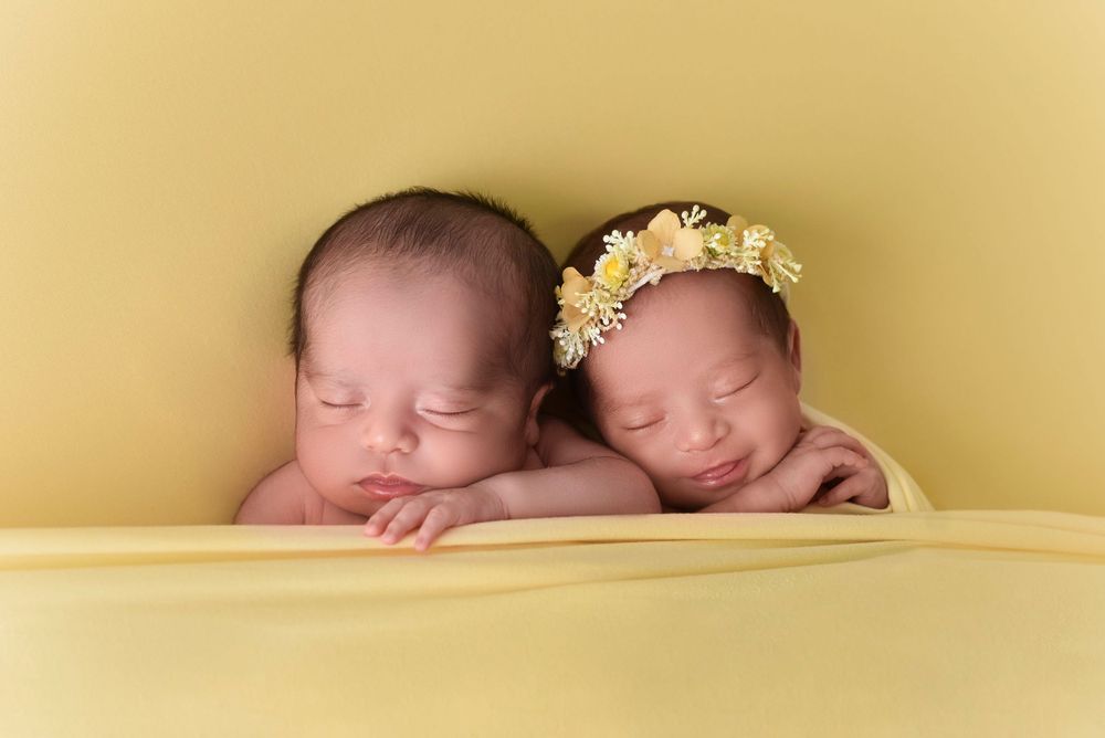 Two sleeping infants wearing delicate flower crowns rest peacefully on a soft yellow background.