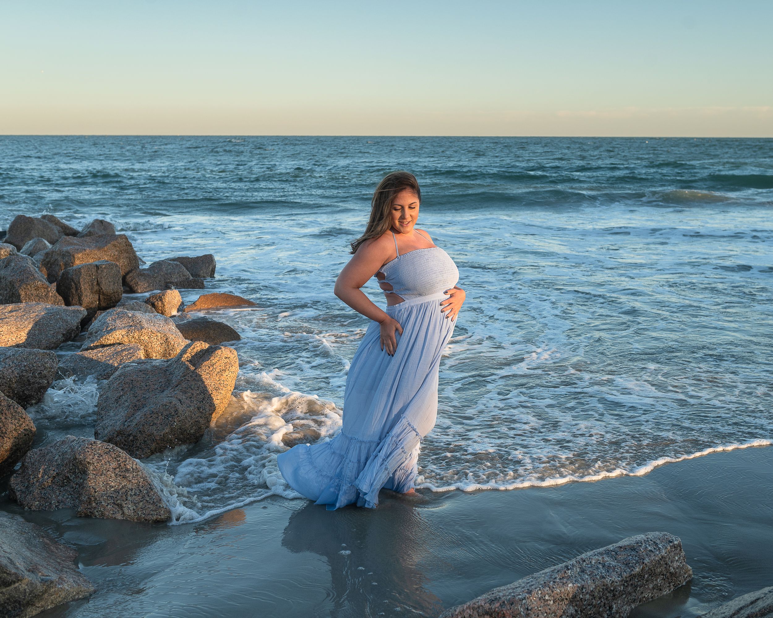 Person in white dress sitting on rocky beach during sunset as waves crash against the shore.
