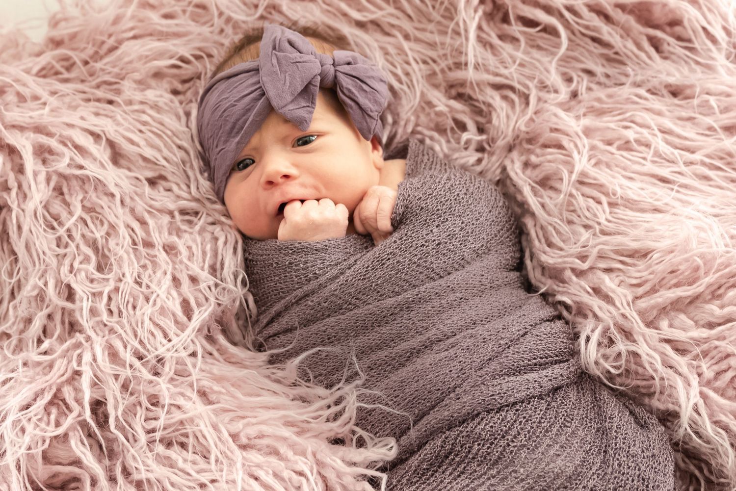 Baby wearing purple headband swaddled in gray wrap on pink fuzzy blanket.