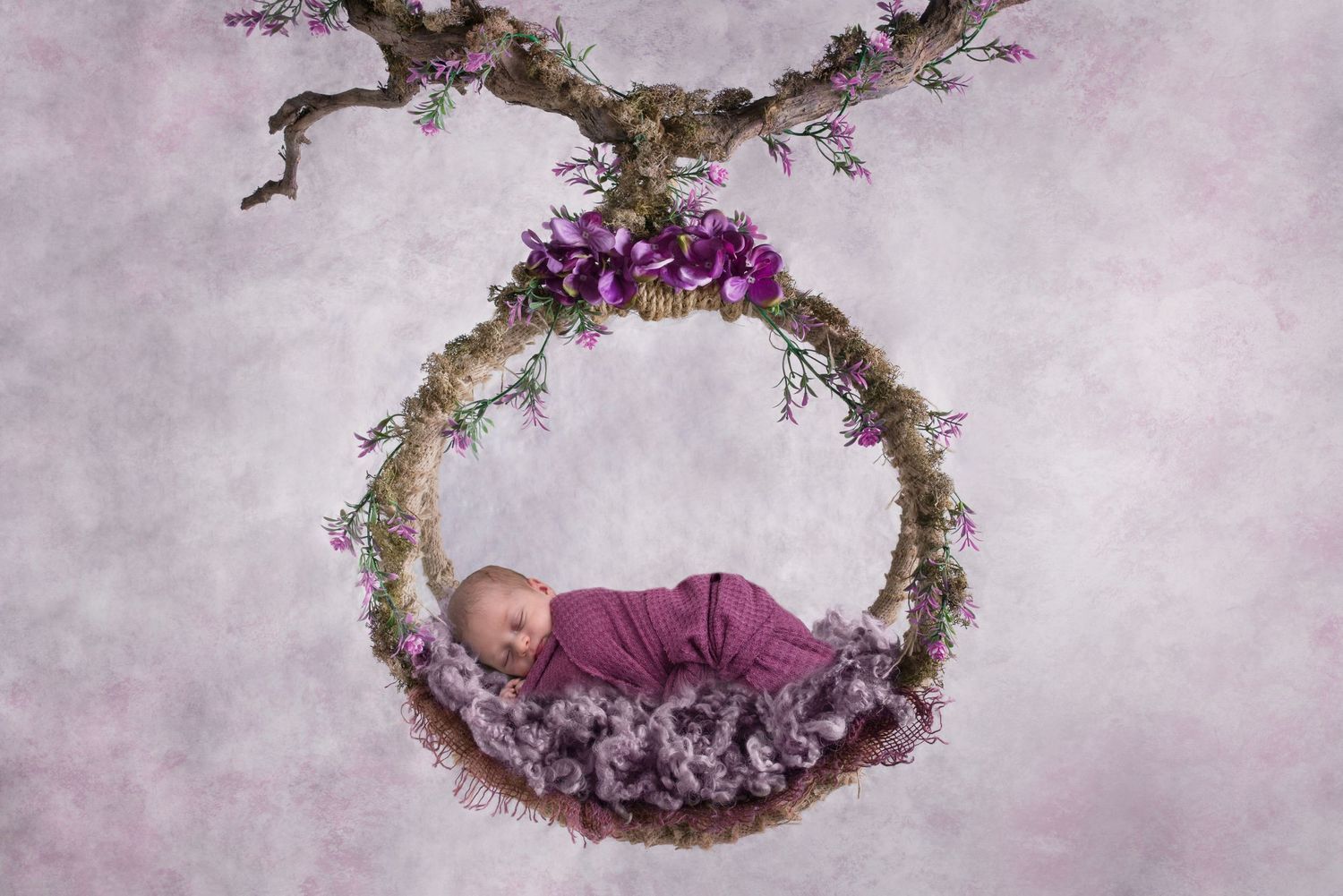 Artistic newborn photo of sleeping baby in purple nest-like arrangement with flowers.