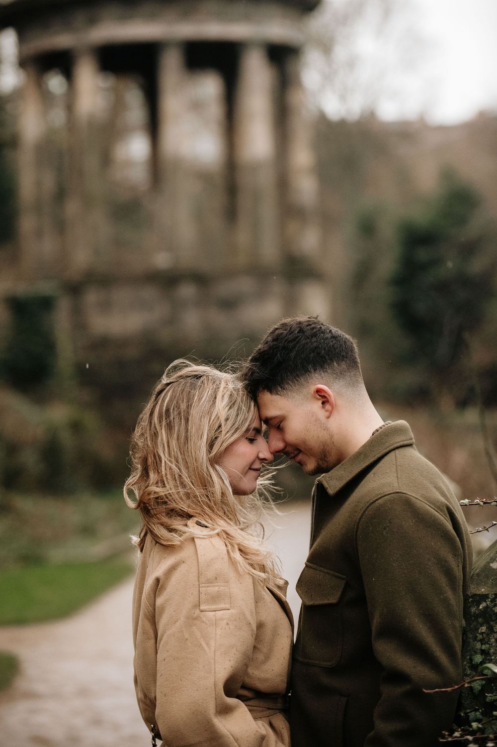 A romantic couple shares an intimate moment on a path with a classical stone structure in the blurred background.